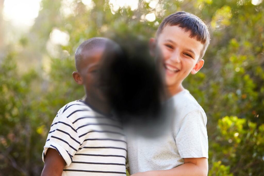 Two young boys laughing in front of a stand of trees. A large black smudge in the center of the photo obscures one boy's face and the other's chest.