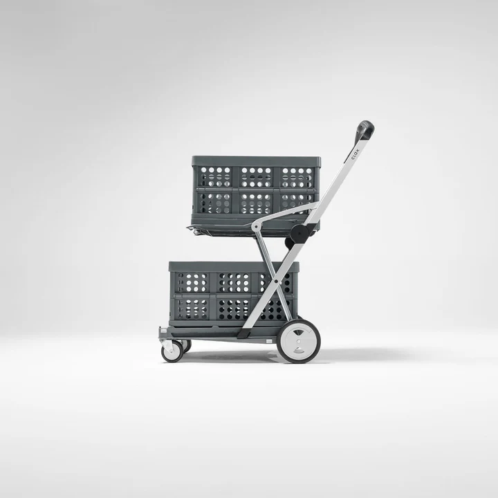 This cart consists of two smaller wheels in the front and two larger wheels in the back, with none of the wheels larger than about six inches. the four wheels are attached to a flat surface. the arm that becomes the handle attached about one third of the way between the back and front wheels and is made of light metal silver in color in this photo. The handle at the top roughly resembles that of a grocery cart (trolly, buggy, pick your term). A second metal arm stretches up from the back wheel, crosses the first arm, and then connects halfway across the second flat surface, which is set back slightly from the first due to the angle of the arms. A third arm connects to the second arm where it met the higher platform and attaches to the first arm just above the upper flat area. in this photo, two grey bins resembling milk crates sit, one on each of the two flat areas. The background of the photo is the white of a photographer's backdrop.